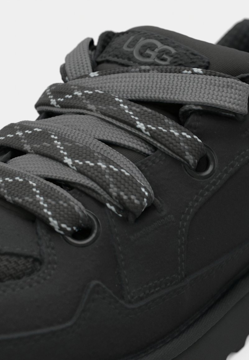 Zapatilla negra con superficie texturizada y detalles en gris. Presenta cordones gris oscuro con un patrón de cuadros y suela de goma.