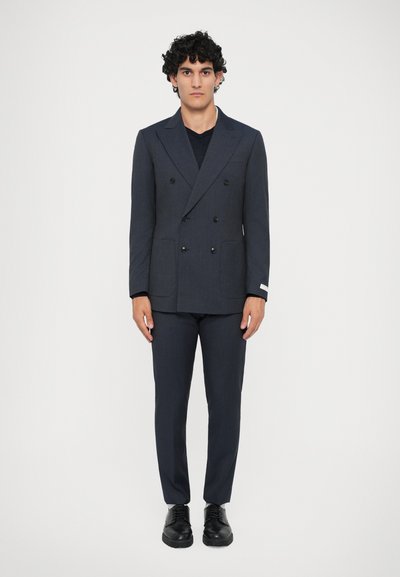 Navy double-breasted suit jacket with black buttons, two front pockets, and matching trousers; paired with a black shirt and shoes.