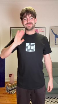 Νέος άνδρας με καστανά μαλλιά και γενειάδα, ντυμένος με μαύρο μπλουζάκι, χαιρετάει μέσα σε εσωτερικό χώρο μπροστά από τοίχο με έργα τέχνης, καναπέ και ξύλινο δάπεδο με σωρούς βιβλίων.