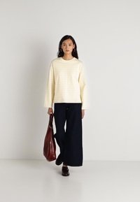 Κρέμα ριγέ μπλούζα με μακριά μανίκια, συνδυασμένη με φαρδιά ναυτικά παντελόνια. Το μοντέλο κρατάει μία σκούρα κόκκινη τσάντα και φοράει καφέ παπούτσια. Μινιμαλιστικό φόντο.