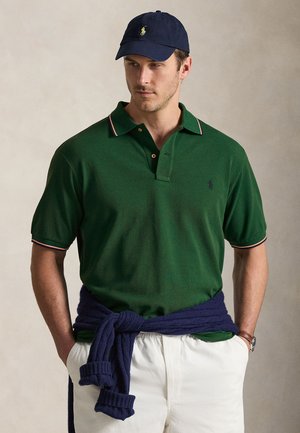 Mężczyzna w ciemnozielonej koszulce polo, granatowej czapce, białych spodniach i granatowym swetrze przewiązanym wokół talii, stoi z rękami w kieszeniach.