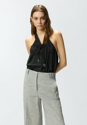 Joven con cabello largo y castaño que lleva un top negro de tirantes con cordones en la parte delantera y pantalones de talle alto color gris claro, de pie frente a un fondo liso.