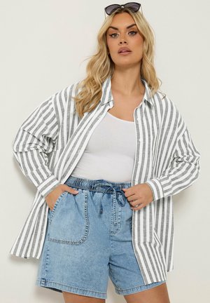 Blond kvinde bærer en grå og hvid stribet skjorte med knapper over en hvid top, kombineret med højtaljede denimshorts og solbriller på hovedet.