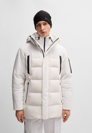 Man in een witte gewatteerde winterjas met zwarte ritsaccenten en een zwarte beanie op een effen lichte achtergrond.