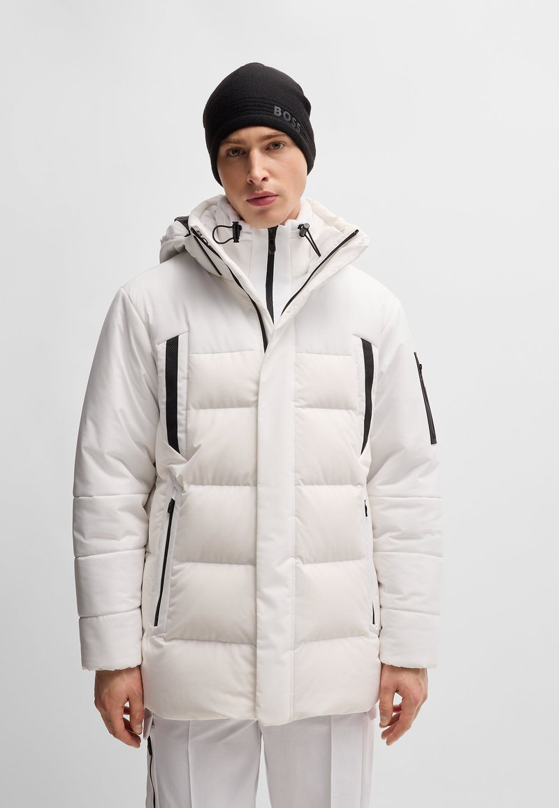 Mann in einer weißen gepolsterten Winterjacke mit schwarzen Reißverschlussakzenten und einer schwarzen Mütze vor einem schlichten hellen Hintergrund.