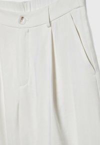 Pantalon blanc taille haute avec passants de ceinture, fermeture avant à boutons et poches latérales, montrant la texture du tissu et les détails des coutures.