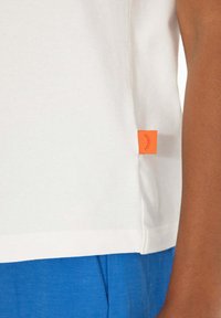 Boční pohled na osobu oblečenou v bílé košili s oranžovým štítkem a jasně modrými kalhotami na jednobarevném pozadí.