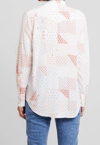 Femme portant une chemise blanche à manches longues avec des motifs en patchwork rouge et un jean bleu, vue de dos sur un fond uni.