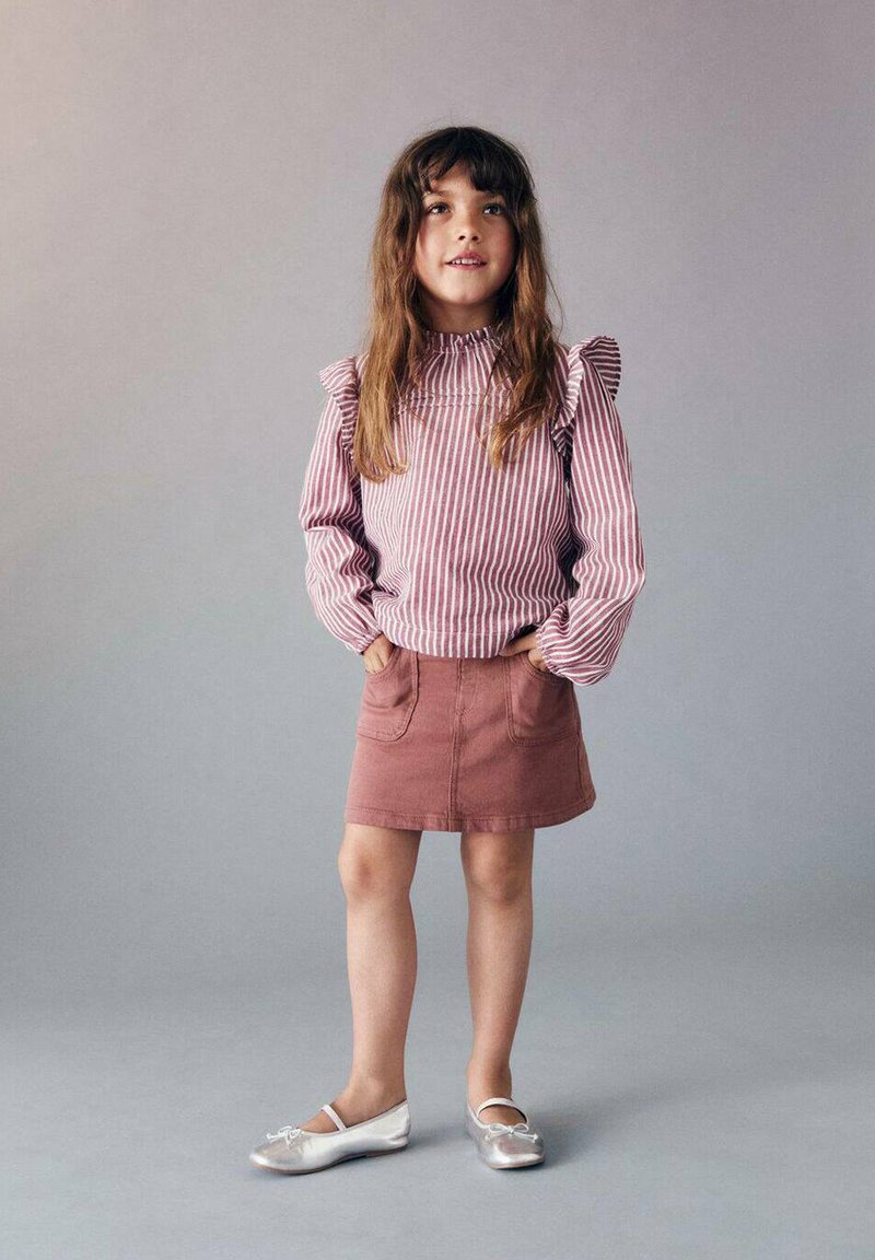 Raidallinen punavalkoinen pitkähihaisten pusero, jossa on röyhelöitä, yhdistetty vaaleanpunaiseen farkkuhameeseen ja hopeisiin ballerinatossuihin. Malli seisoo kädet taskuissa.