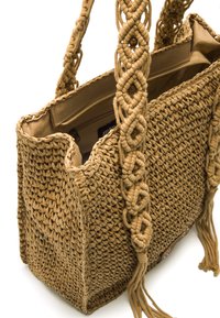 Bolsa tote de paja trenzada en marrón con un acabado texturizado, que cuenta con un asa anudada y borlas laterales. El interior incluye un bolsillo con cremallera.