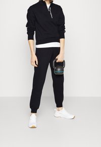 Svart zip-up sweatshirt med vitt logotyp, i kombination med svarta joggers med uppvikta mudd, och vita sneakers med bruna sulor; håller en svart kettlebell.