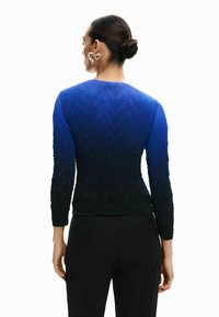 Blaues, langärmliges Strickoberteil mit strukturiertem Zickzackmuster, das unten zu Dunkelblau/Schwarz übergeht, kombiniert mit schwarzen Hosen.