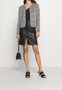 Blazer de cuadros en blanco y negro con cuello redondeado, combinado con una falda lápiz de cuero negro con una abertura frontal. Los accesorios incluyen un pequeño bolso negro.