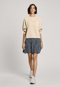 Beige-huppari, jossa on raglan-hihat, yhdistetty siniseen kukkakuosiseksi hameeksi, jossa on levenevä helma. Mallilla on päällään vaaleat lenkkarit.