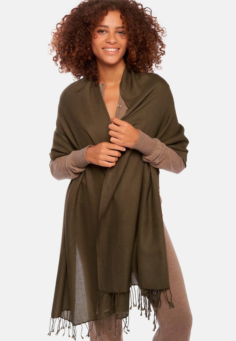 Châle vert olive avec des bords à franges, fait en matériau doux, drapé sur un modèle portant un top à manches longues dans une teinte plus claire.