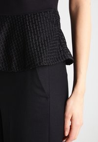 Černý top s texturovaným peplum designem a tkaným vzorem, spárovaný s hladkými černými kalhotami. Materiál má matný povrch.