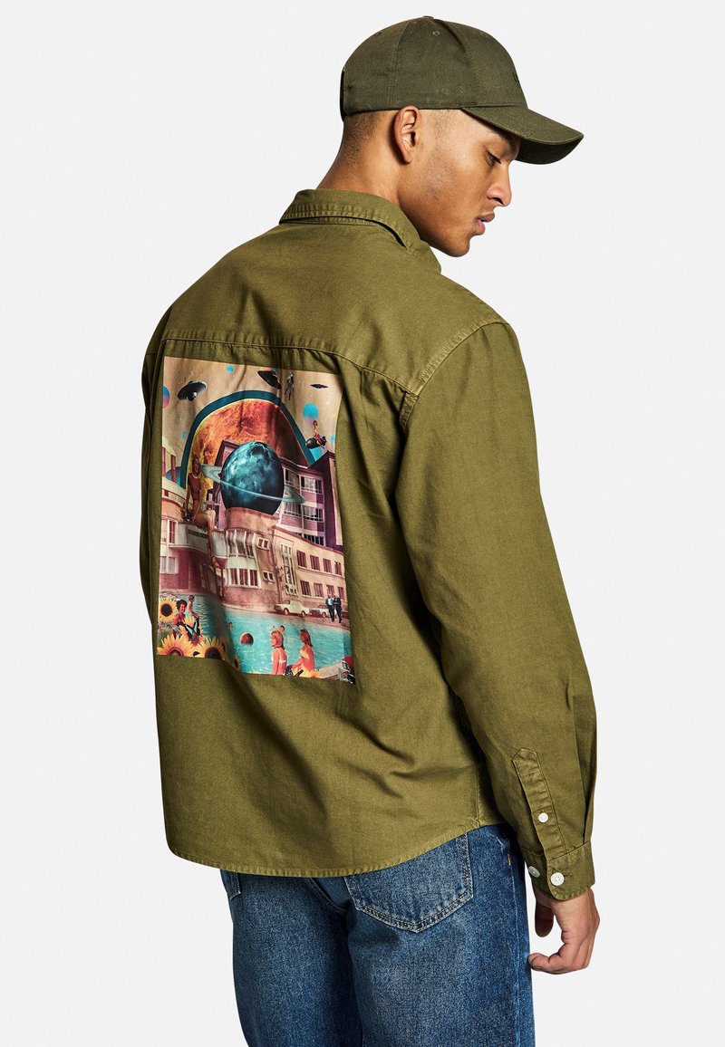 Chemise vert olive à manches longues avec un poignet boutonné, ornée d'un grand graphique coloré au dos représentant un paysage surréaliste.