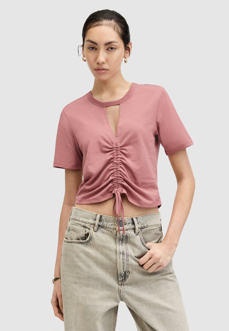 Frau in einem rosa kurzen, gerafften Crop-Top mit einem Schlüssel-Ausschnitt vorne, kombiniert mit hellen, hochgeschnittenen Jeans, steht vor einem grauen Hintergrund.
