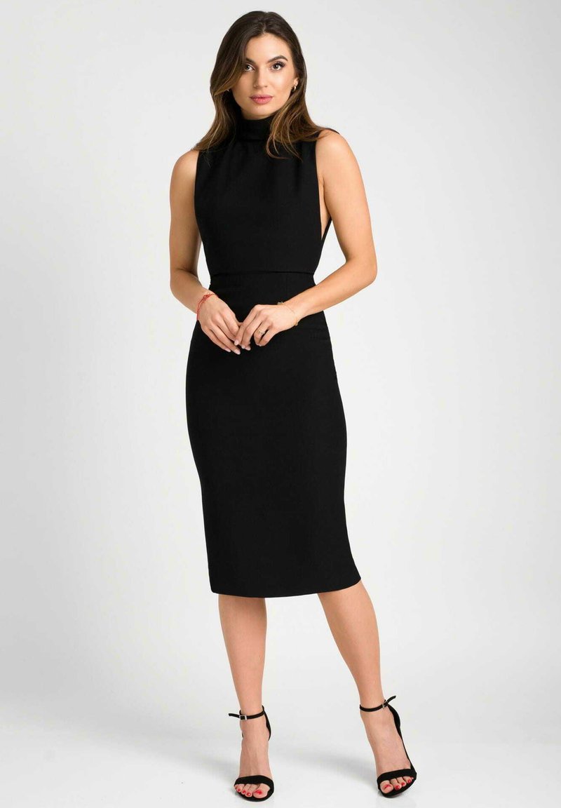 Czarna sukienka do kolan bez rękawów z wysokim dekoltem, dopasowanym krojem i gładkiego materiału. W połączeniu z czarnymi szpilkami na kostkę.