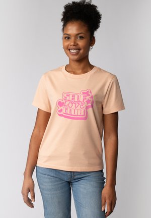 Lächelnde Frau mit lockigem Haar, die ein pfirsichfarbenes T-Shirt mit der Aufschrift "SELF LOVE CLUB" in Pink trägt, kombiniert mit blauer Jeans vor neutralem Hintergrund.