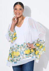 Weißes, transparentes Oberteil mit Glockenärmeln, das einen bunten Zitronen- und Blumenprint am Saum aufweist. Kombiniert mit blauer Jeans.