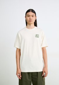 Cream-colored cotton t-shirt with short sleeves and a front green graphic design. Round neckline and relaxed fit. No visible patterns.