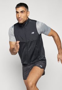 Gilet de sport noir et gris avec une fermeture éclair à l'avant, une section inférieure texturée et un logo réfléchissant. Porté par-dessus un t-shirt gris à manches courtes et un short à motifs.