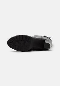 Bota de cuero negro tumbada de lado mostrando suela de goma texturada y tacón cuadrado sobre un fondo blanco.