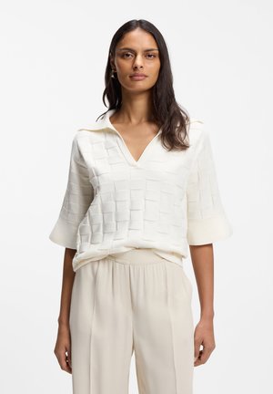 Γυναίκα με μακριά σκούρα μαλλιά που φοράει λευκό υφασμάτινο κοντομάνικο τοπ με γιακά και ανοιχτό μπεζ παντελόνι ψηλόμεσο με φαρδιά μάνικα, στεκόμενη μπροστά σε λευκό φόντο.