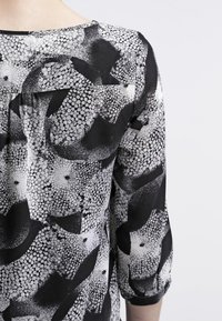 Blouse à motifs en noir et blanc avec un tissu doux, présentant des formes et textures abstraites. Manches trois-quarts avec un col arrondi.