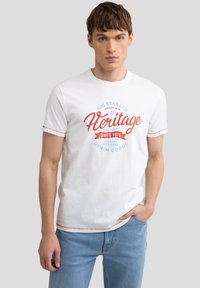 Biała bawełniana koszulka z kolorowym nadrukiem "Heritage", z krótkimi rękawami i kontrastowym przeszyciem, w luźnym kroju. W zestawieniu ze jasnoniebieskimi dżinsami.