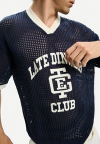 Maglione a rete blu marina con colletto a V bianco e testo "LATE DINNER CLUB" in lettere grasse, che presenta un design morbido e traspirante dallo stile sportivo.