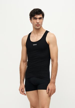 Homme portant un débardeur noir Versace et un boxer noir, debout devant un fond clair uni.