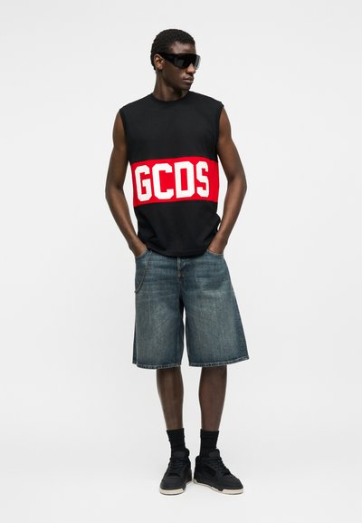 Άνδρας που φοράει μαύρο αμάνικο κορμάκι με κόκκινο και λευκό λογότυπο GCDS, τζιν σορτς μέχρι το γόνατο, μαύρα αθλητικά παπούτσια, μαύρες κάλτσες και μεγάλα μαύρα γυαλιά ηλίου.