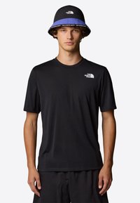 Camiseta negra de manga corta con cuello redondo y pequeño logotipo blanco en el lado izquierdo del pecho, combinada con un sombrero tipo cubo con acentos azules y detalles de la marca.