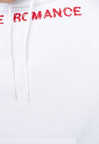 Vit hoodie med röd broderad text "LOVE ROMANCE", försedd med justerbara dragskoar och mjukt material.