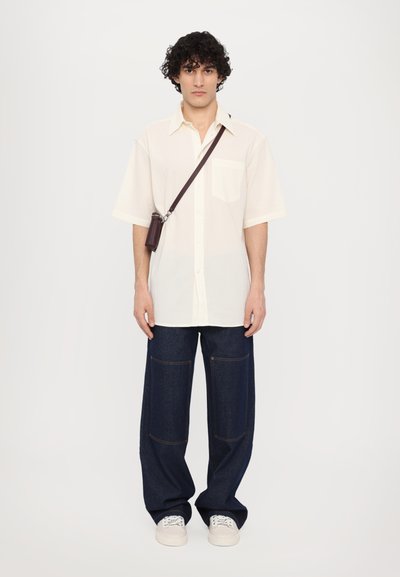 Chemise créme à manches courtes avec une poche, associée à un jean en denim bleu foncé à jambes larges avec deux poches avant et des baskets blanches.