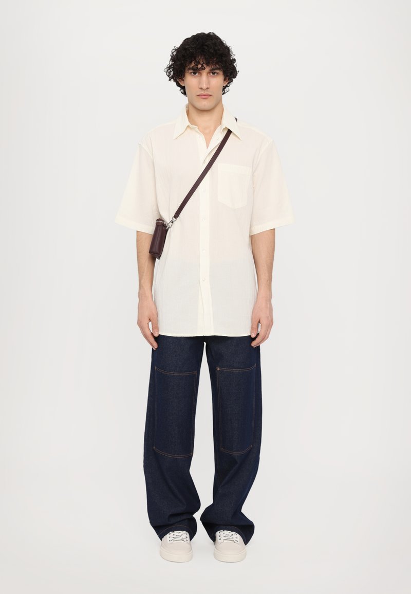 Camisa crema de manga corta con un bolsillo, combinada con pantalones vaqueros de pierna ancha azul oscuro que cuentan con dos bolsillos frontales y zapatillas blancas.