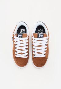 Ténis de camurça castanhos com atacadores brancos, colarinho acolchoado e forro interior preto, apresentando um pequeno logótipo na língua e na lateral.