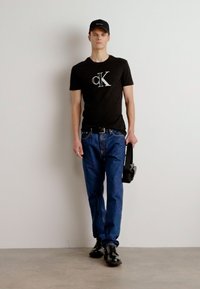 Must musta T-särk logoga, sinised teksapüksid, mustad kingad ja must müts; inimene hoiab musta kotti, seistes neutraalse tausta ees.