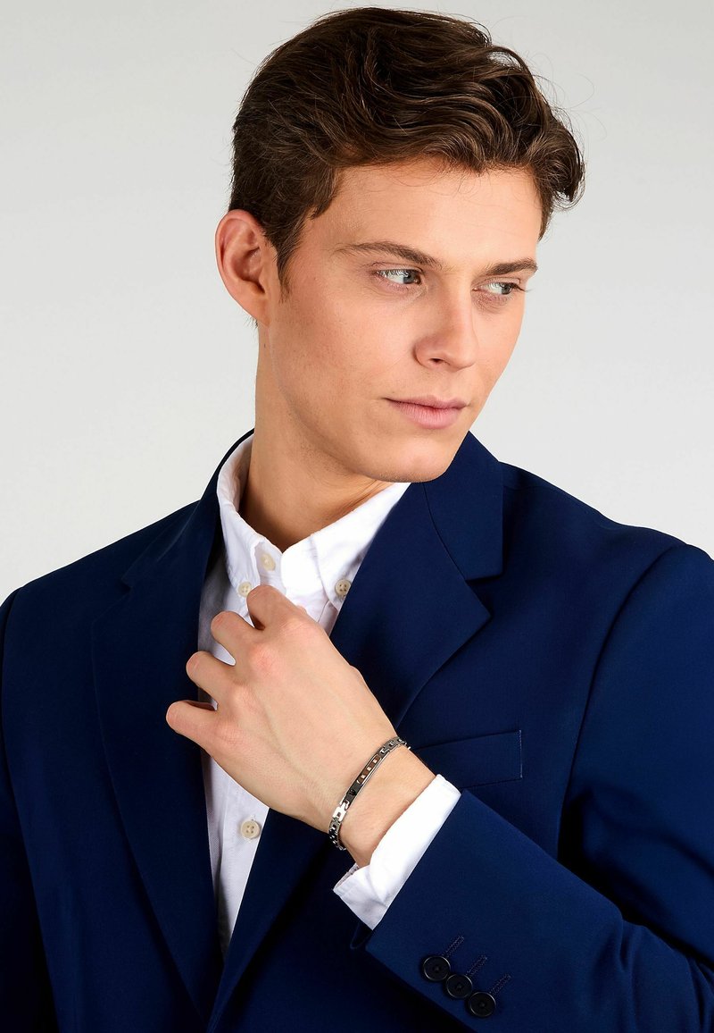 Bracciale d'argento con maglie rettangolari, indossato su una mano che sistema una camicia bianca con colletto sotto un blazer blu scuro.