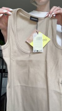 Béžový žebrovaný top bez rukávů s značkovou cedulkou "even&odd" a přiloženými informacemi o produktu, který drží osoba s upravenými nehty.