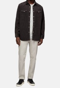 Mężczyzna w ciemnoniebieskiej koszuli z podwijanymi rękawami na białym t-shircie, jasnych beżowych spodniach i czarnych trampkach z białymi podeszwami, stojący.