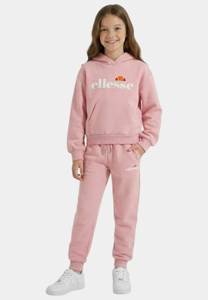 Jeune fille souriante portant un sweat à capuche ellesse rose et un pantalon de survêtement assorti avec des baskets blanches, debout avec une main dans la poche.
