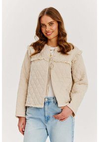 Beżowa pikowana kurtka z falbankowym kołnierzem i wiązaniem. Posiada długie rękawy oraz fakturowany materiał. Noszona na białej bluzce.