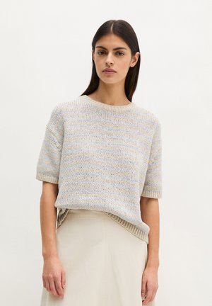 Femme aux cheveux foncés portant un pull en maille à manches courtes rayé beige et bleu clair ainsi qu'une jupe couleur crème, sur un fond uni.