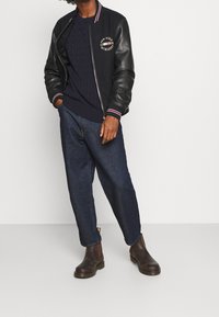 Giacca varsity in pelle nera e lana blu navy con colletto a righe rosse e bianche, indossata su un maglione blu navy e jeans scuri a taglio ampio, abbinata a stivali alla caviglia marroni.