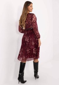 Bordeauxrotes plissiertes Kleid mit floralem Muster, langen transparenten Ärmeln und schwarzem Gürtel in der Taille; kombiniert mit hohen schwarzen Stiefeln mit Absatz.