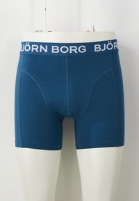 Kék pamut boxeralsó fehér elasztikus derékkal, amelyen a "BJÖRN BORG" felirat olvasható. Sima textúrájú, testhezálló fazonú és combközép hossza van.