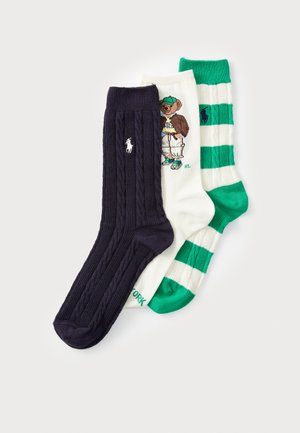 Trois paires de chaussettes : en tricot torsadé bleu marine, blanches avec des rayures vertes, et blanches avec un graphique de ours. Toutes ont des logos de marque contrastants.